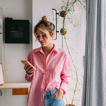 An individual stands indoors holding a smartphone in one hand, with plants, shelving, and window curtains visible in the background.